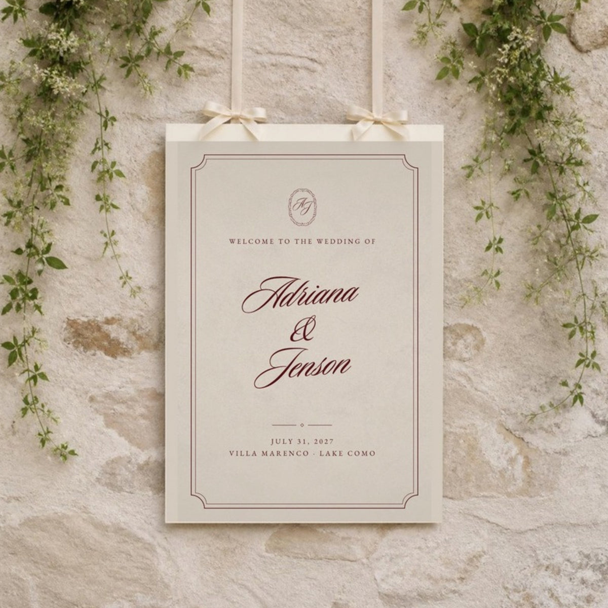 Ivory welcome sign hanging from ribbons on a stone wall with greenery